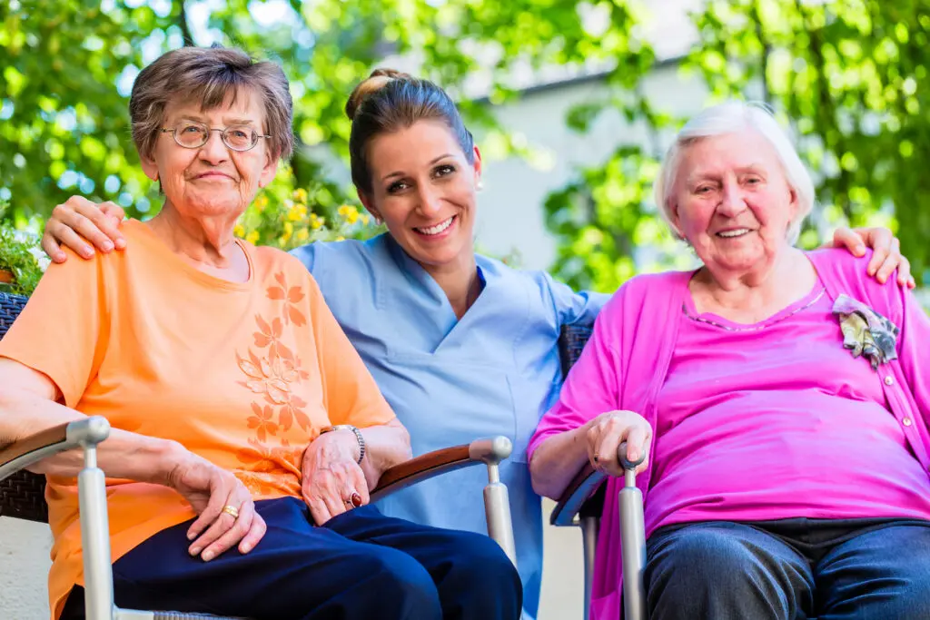 Three women sitting in wheelchairs with a nurse standing behind them, surrounded by trees and a building in the background