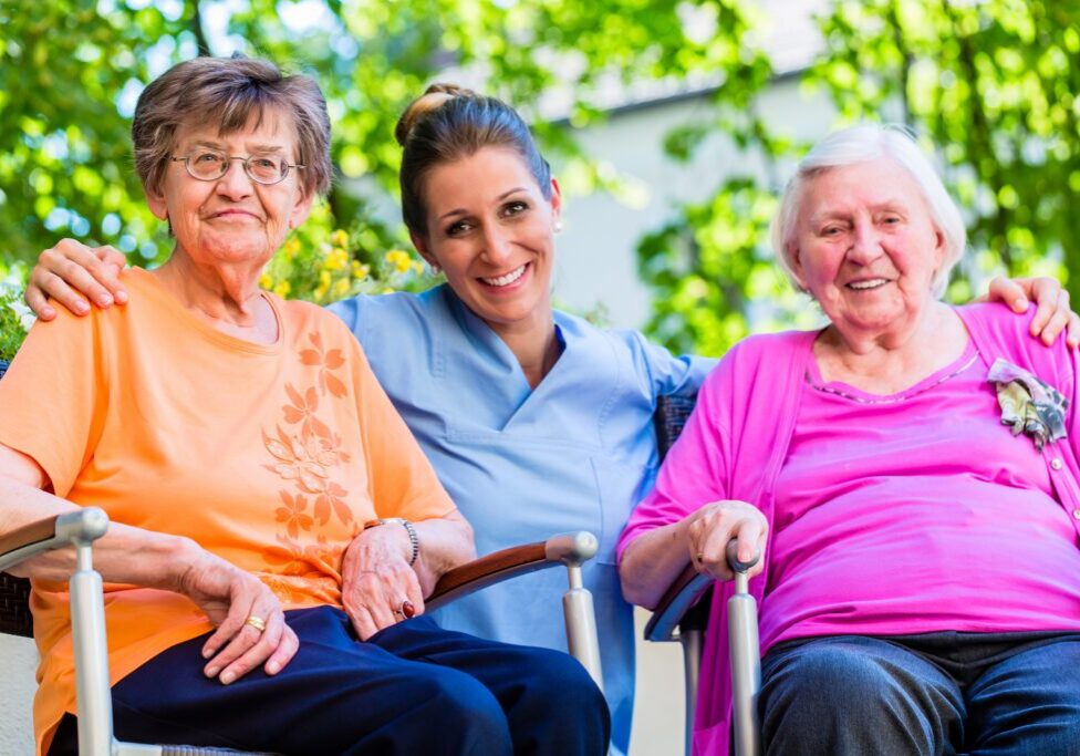 Three women sitting in wheelchairs with a nurse standing behind them, surrounded by trees and a building in the background
